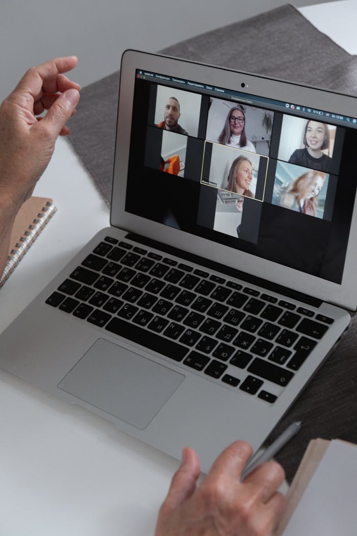 Hands at a laptop showing a group video call in a home office setting.